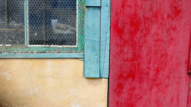 A photo of a red and beige farm wall in northern France. There’s a blue window frame with chicken-wire over it